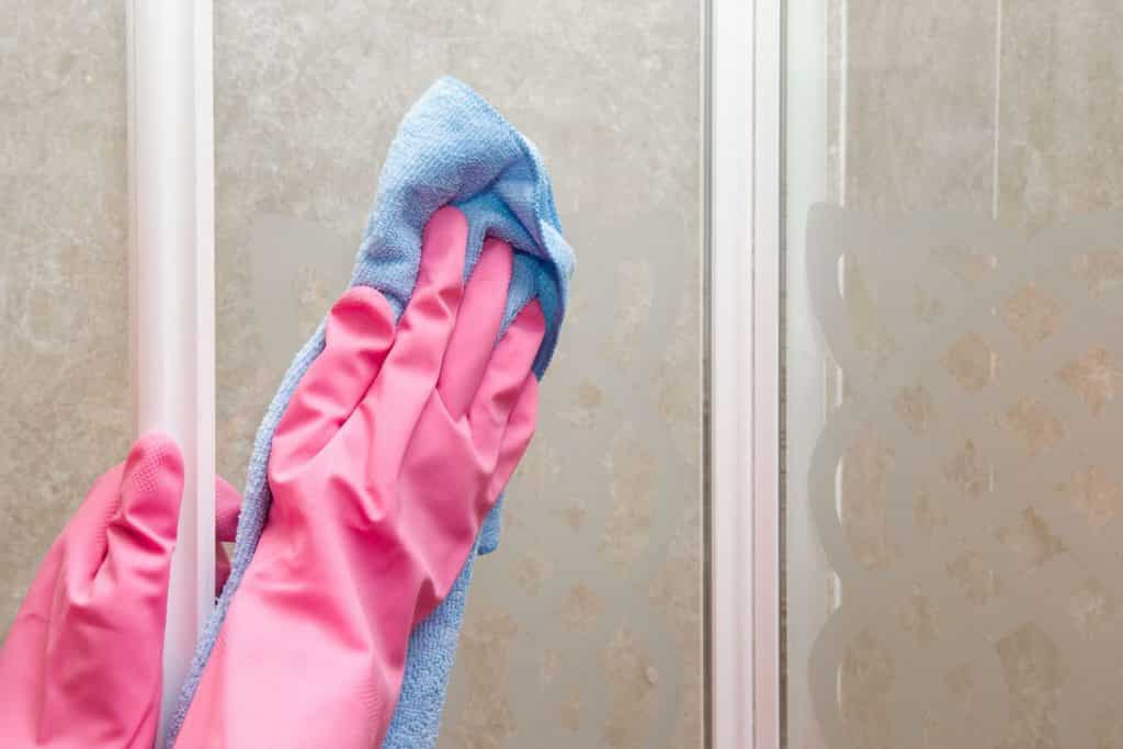 A professional house cleaner wiping a glass shower wall with a microfiber cloth.